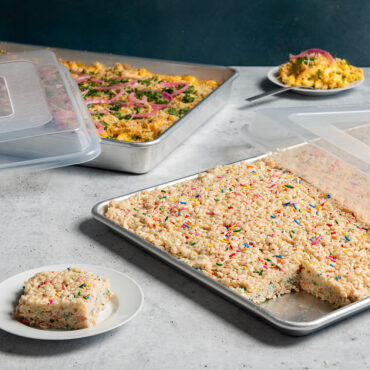 Two Nordic Ware aluminum baking pans with clear plastic storage lids, one containing baked mac and cheese, the other with rice crispy treats