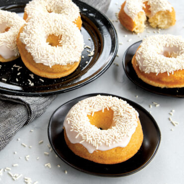 Six glazed vanilla donuts with white sprinkles arranged on white plates, baked in Nordic Ware red donut pan