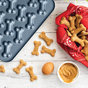Nordic Ware blue aluminum Puppy Love Treat Pan with bone-shaped cavities, alongside baked golden dog treats