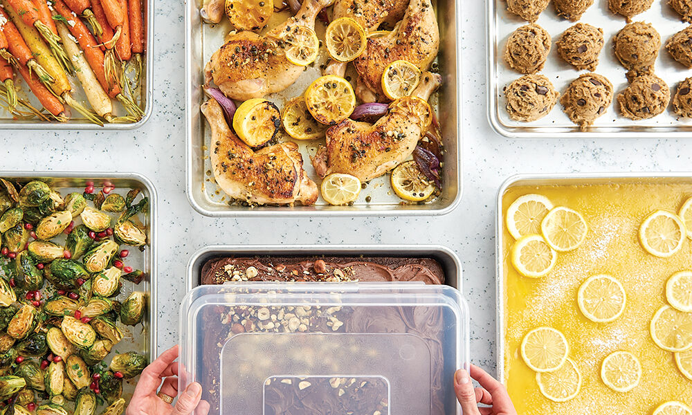 Six Nordic Ware Naturals Half Sheet Pans and High Sided Half Sheet Pans. One is filled with cookie dough balls, one has lemon bars, one has chicken thighs with lemon, one has a chocolate hazelnut sheet cake, one has roasted carrots, and one has roasted brussels sprouts. There are two hands placing a Half Sheet Lid on top of the pan with the chocolate hazelnut cake in it.