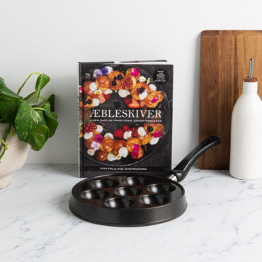 Nordic Ware cast aluminum aebleskiver pan with seven round wells and accompanying cookbook on marble surface