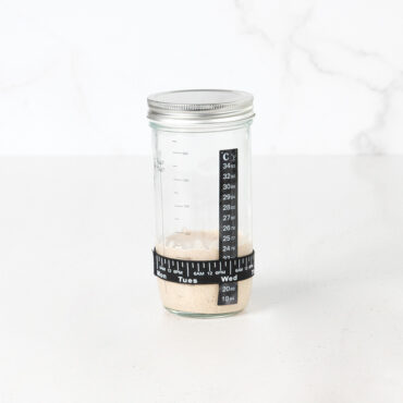 Glass starter jar with bubbly sourdough starter inside, sitting on white kitchen counter