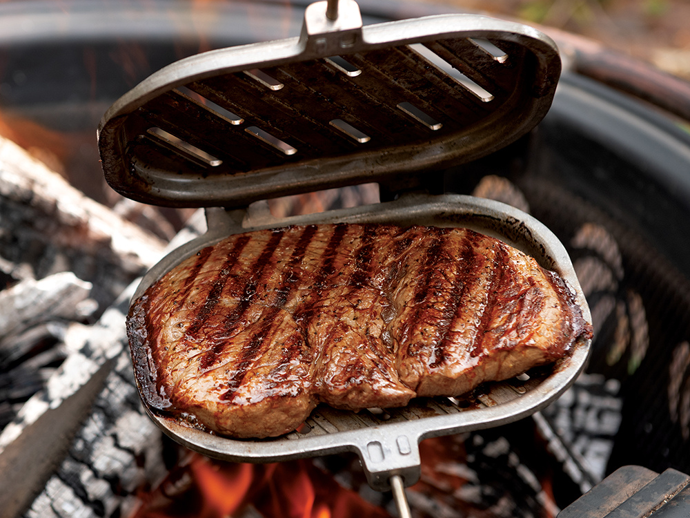 Campfire Griller open over a bonfire with a large steak cooking in it. You can see steak grill lines left from the open flame on the steak.