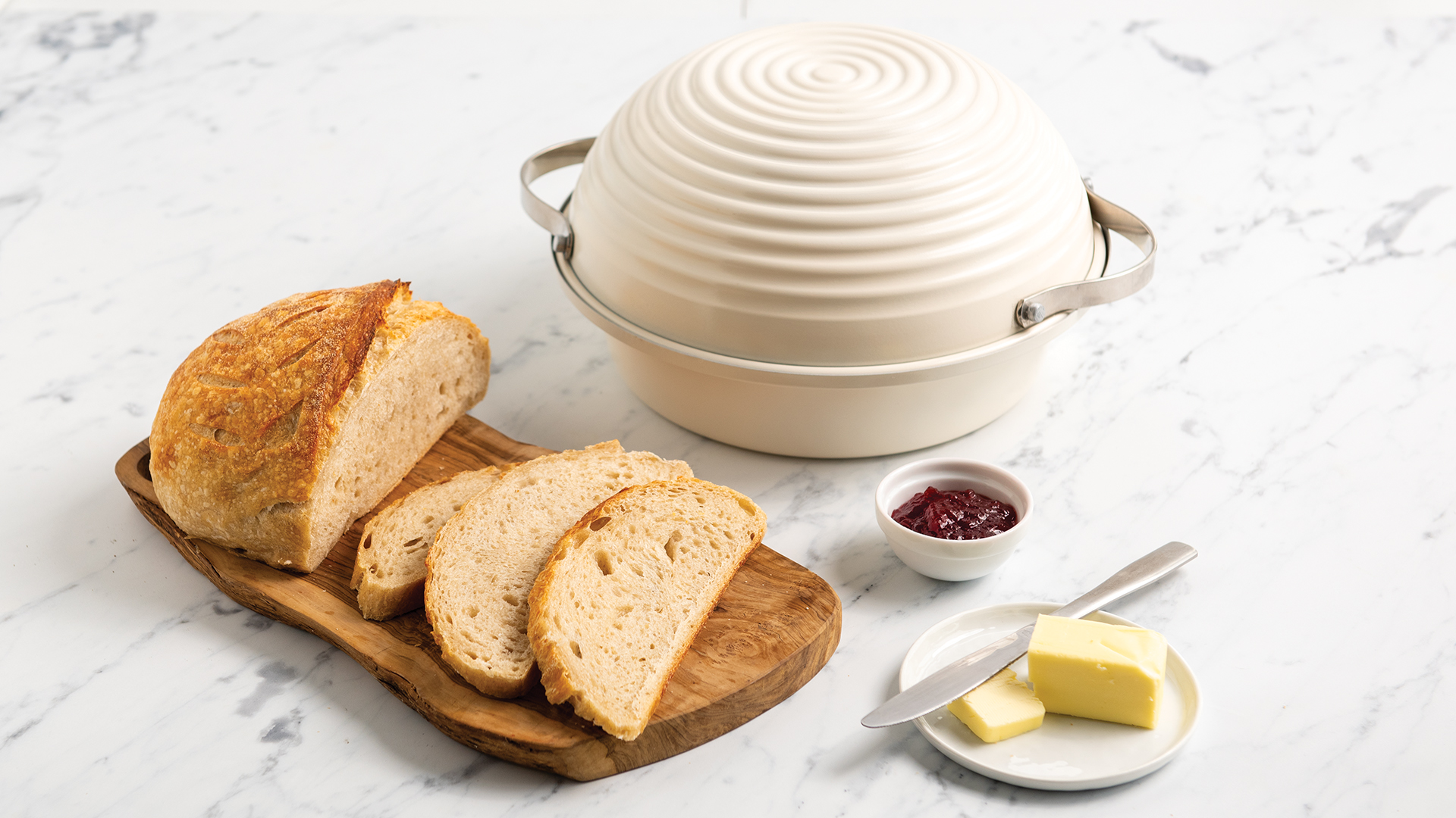 A perfectly baked loaf of sourdough bread sliced and served on a wooden cutting board with butter and jam next to the Simply Sourdough Bread Baking Pan. 