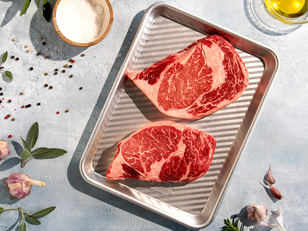 3-in-1 Prep, Grill and Serve Tray with two large raw steaks on it. Surrounding the tray, there are various seasonings, garlic, and olive oil.