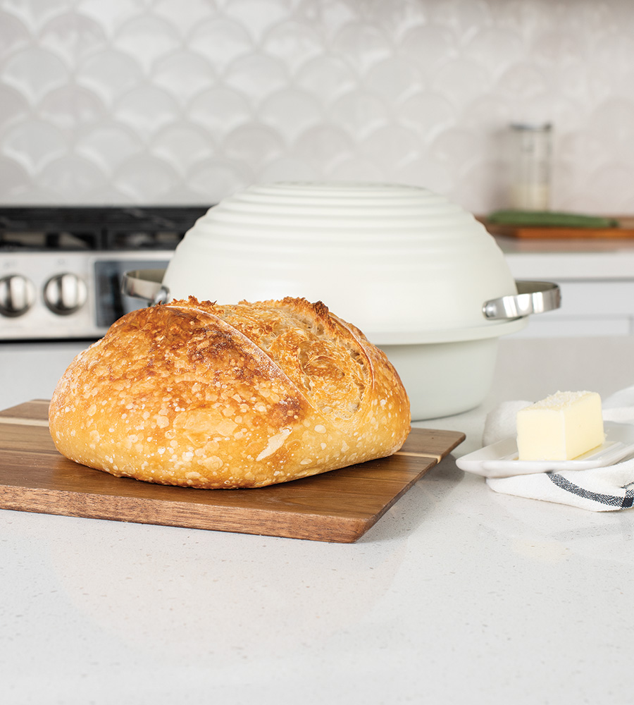 Baked sourdough bread loaf on a cutting board in a kitchen with Nordic Ware baking pan behind it.