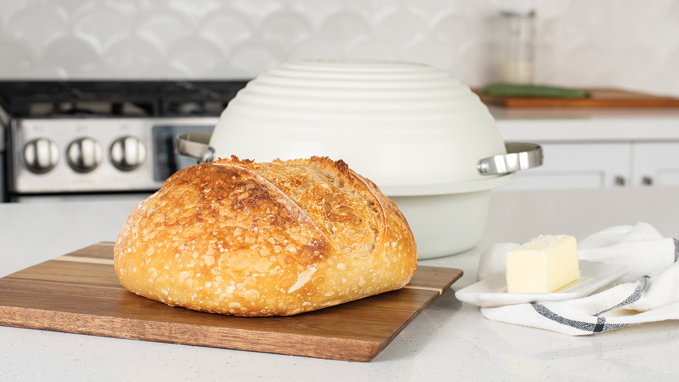 Image of baked sourdough bread loaf on a cutting board in a kitchen with Nordic Ware baking pan behind it.