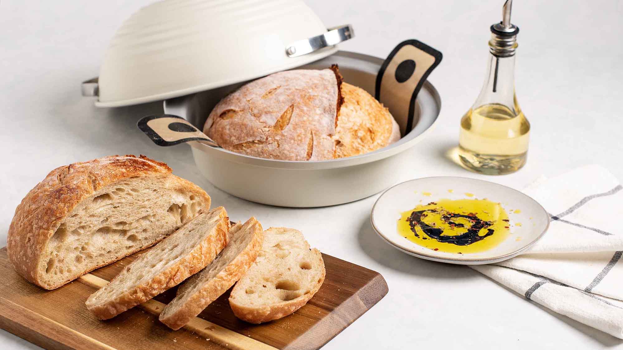 A baked loaf of sourdough bread sitting inside of the Simply Sourdough Bread Baking Pan next to a sliced loaf of sourdough bread on a wooden cutting board that is ready to be served.