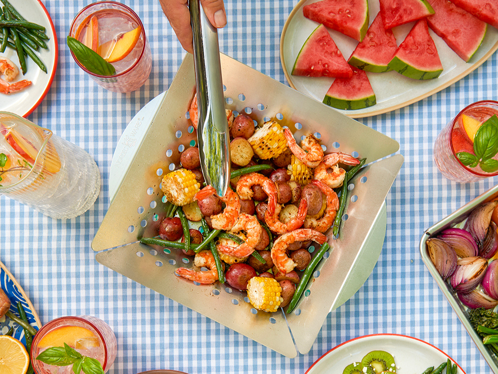 Perforated Grilling Basket serving grilled vegetables and shrimp on a blue checkered tablecloth. A hand holding tongs is serving the food. On the table, there is also a plate of watermelon, three full drinks, and plates with the serves shrimp and vegetables on them.