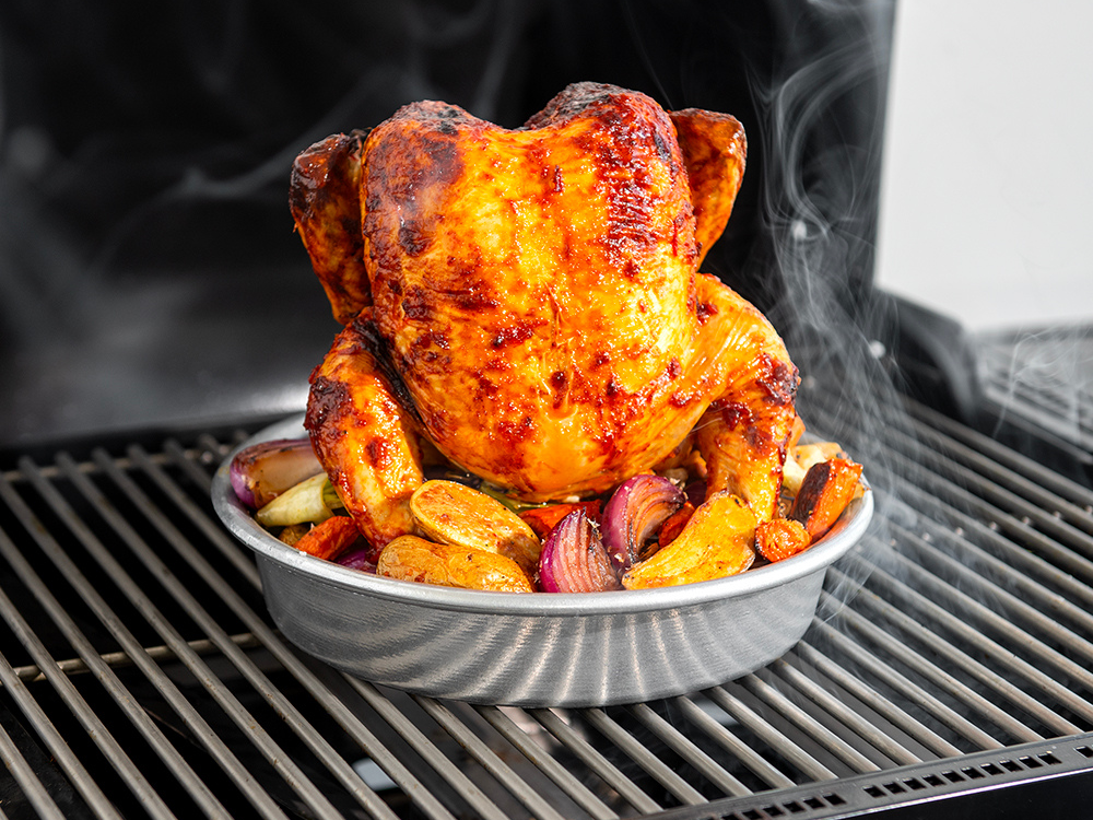Vertical Chicken Roaster on the grill cooking a chicken to golden, crispy perfection. In the base of the Vertical Chicken Roaster are vegetables that are cooking in the drippings of the chicken.
