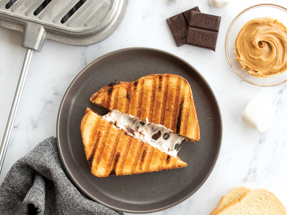 Grilled Peanut Butter S’mores Sandwich served on a gray plate. The sandwich is cut in half and gooey marshmallow is oozing out of the sandwich. Next to the sandwich, there is the Campfire Griller, that was used to cook the sandwich, along with chocolate, marshmallows, peanut butter, and bread on the table ready to make another sandwich.