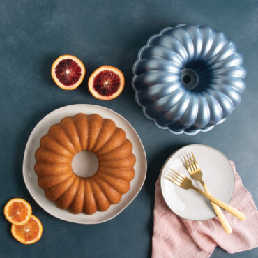 Golden bundt cake on white plate beside Nordic Ware Elegant Party bundt pan with orange and mango slices, two gold forks on empty plate