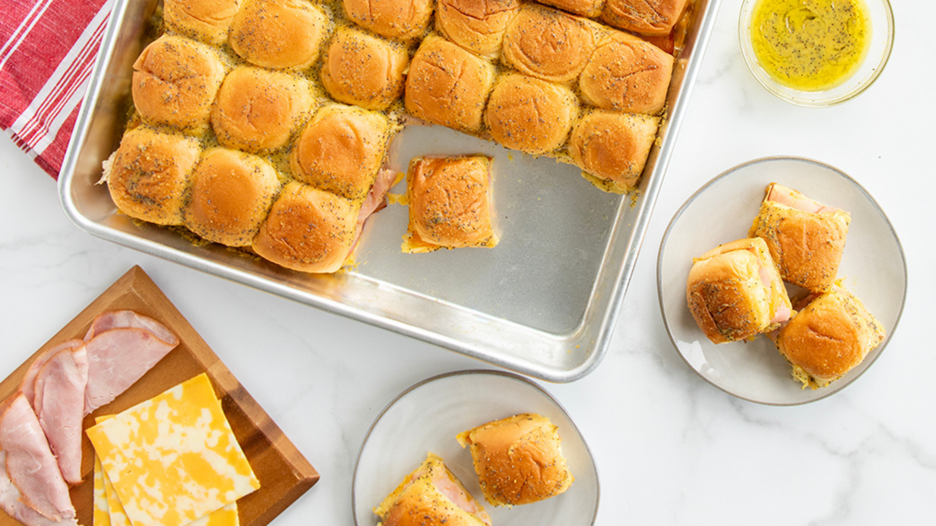 Baked Ham and Cheese Sliders in the Naturals® Big Batch Pan. Some sliders are served on plates next to the pan along with some additional ham, cheese, and butter on the side.