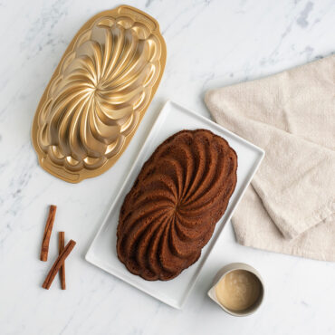 Pirouette Loaf cake on plate next to pan, cup of frosting, and cinnamon sticks