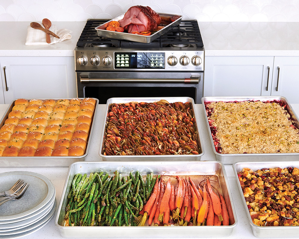 Big Batch Baking Pans filled with food for a holiday meal including bread rolls, stuffing, vegetables, ham, and crumble.
