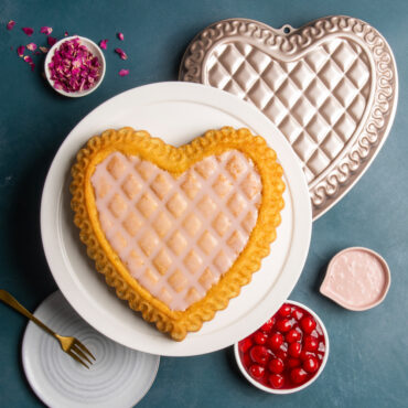 Overhead of quilted heart cake with icing on white cake stand with Pan, petals, dish of cherries, and pink icing around