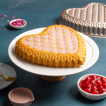 Uuilted heart cake with icing on white cake stand with Pan, petals, dish of cherries, and pink icing around