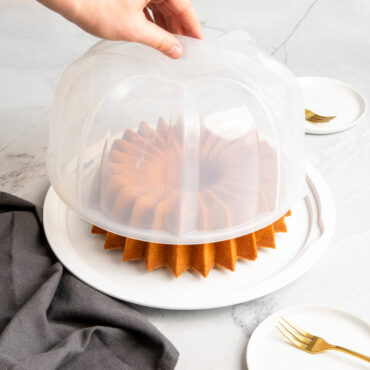 Clear plastic bundt cake keeper with domed lid containing golden baked brilliant bundt cake, marble background, hand holding lid over base