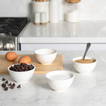 Four white mini bowls arranged on white countertop with chocolate chips and brown sugar, cutting board with eggs on it