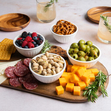 Four mini white bowls on a charcuterie board filled with snacks and cheese shot at an angle