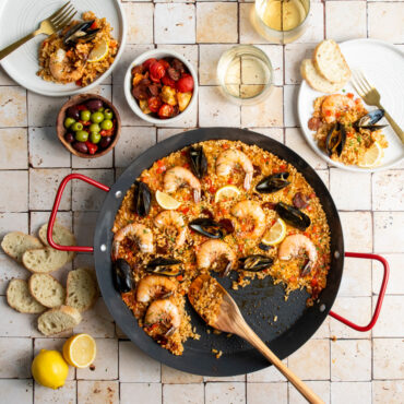 paella pan with saffron rice, shrimp, mussels, and vegetables displayed on tile countertop, wooden spoon in pan with two plated portions