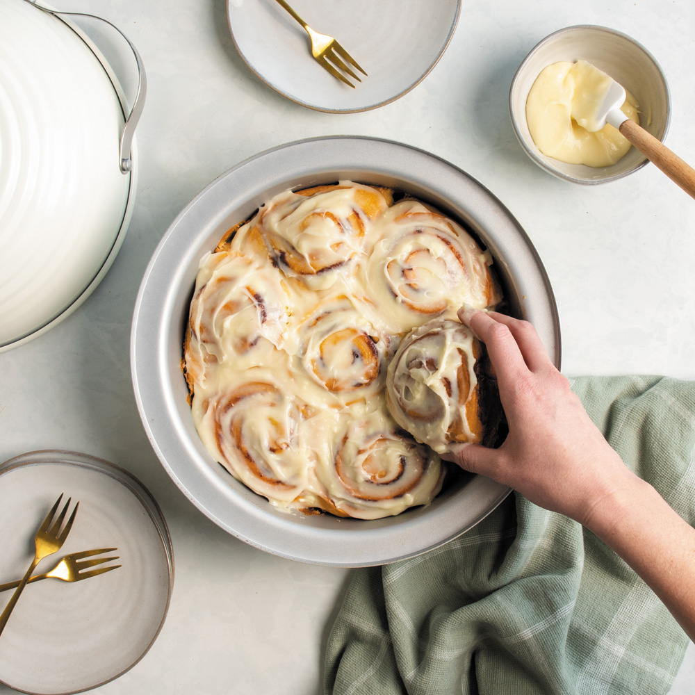 Image of baked sourdough cinnamon rolls in Nordic Ware Sourdough Bread Baker Pan with hand grabbing a single cinnamon roll.