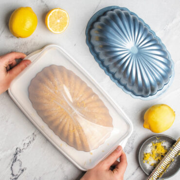 overhead image of lemon fluted loaf cake in keeper on marble countertop, hands closing lid, lemons and pan scattered around