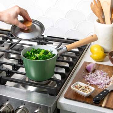 cardamom 1.5qt saucepan on stovetop with broccoli, hand holding lid above pan