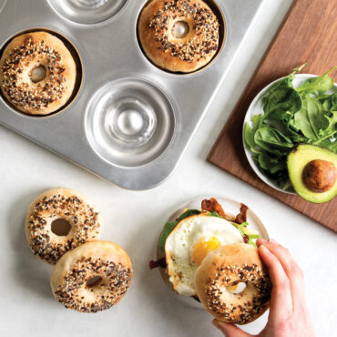 overhead of sourdough bagel pan with bagels in pan, hand placing bagel half on plated egg sandwich