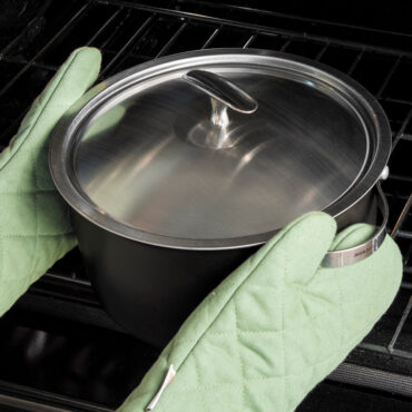 hands wearing oven mitts pulling nordic ware 6 qt aluminum dutch oven and lid with stainless steel handles and stainless steel lid and knob out of the oven