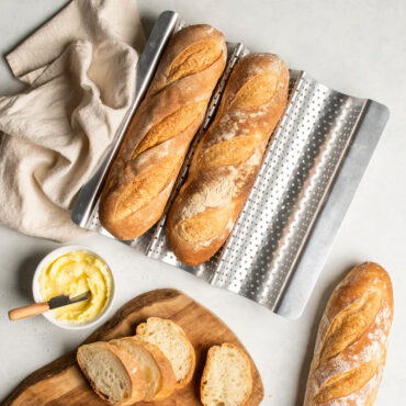 two fresh baked baguettes nested in a well of Nordic Ware baguette & french bread pan; one baguette cut into slices on cutting board