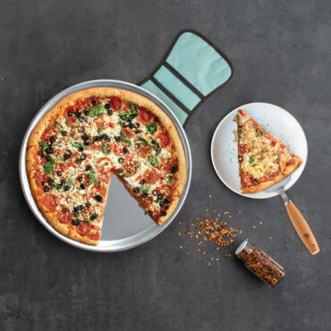 nordic ware deep dish pizza pan with supreme pizza, hot pad underneath pizza pan, plated slice of pizza rested on caker server and plate