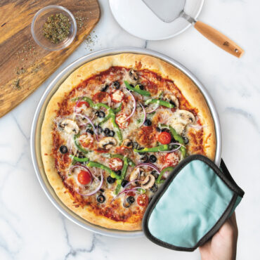 hand with hot pad holding baked pizza in nordic ware deep dish pizza pan, cake server resting on white plate