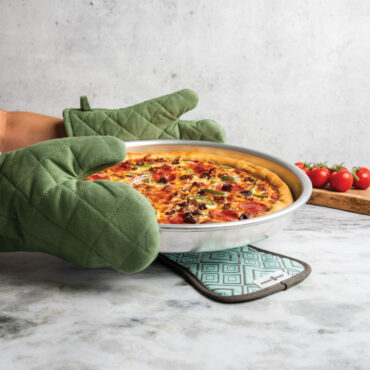 hands with oven mitts holding nordic ware 14 inch deep dish pizza pan with pizza above cotton hot pad with silicone geometric detailing